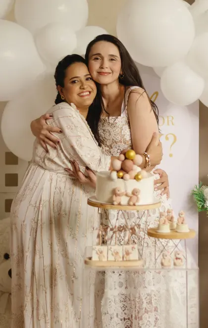 Two women share a warm, tearful embrace before a soft cream balloon arch.
