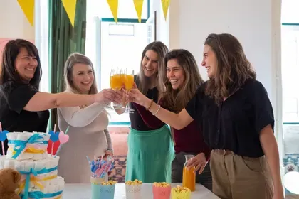 Four friends raise glasses in a cheerful toast beside a diaper cake in a sunlit room.