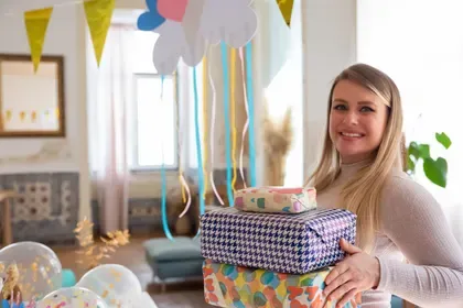 A smiling mom-to-be holds a stack of wrapped gifts under pastel bunting and clouds.