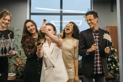 Coworkers in party glasses laugh as a colleague raises a bottle of champagne at the office party.