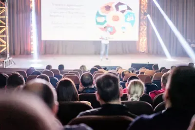 An audience watches a presenter on a bright stage with dramatic light beams and a big screen.