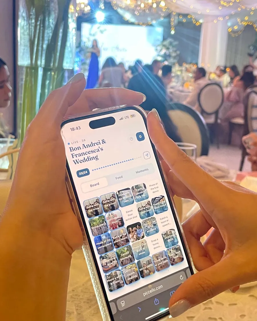 A guest holds a phone showing a Piccello board at a wedding reception, twenty of twenty-four tiles filled with photos, other guests seated at tables behind them.