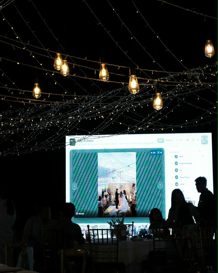 An LED wall at an outdoor evening wedding shows the live Piccello photo feed and leaderboard beneath strings of warm lights, guests seated in silhouette in front of it.